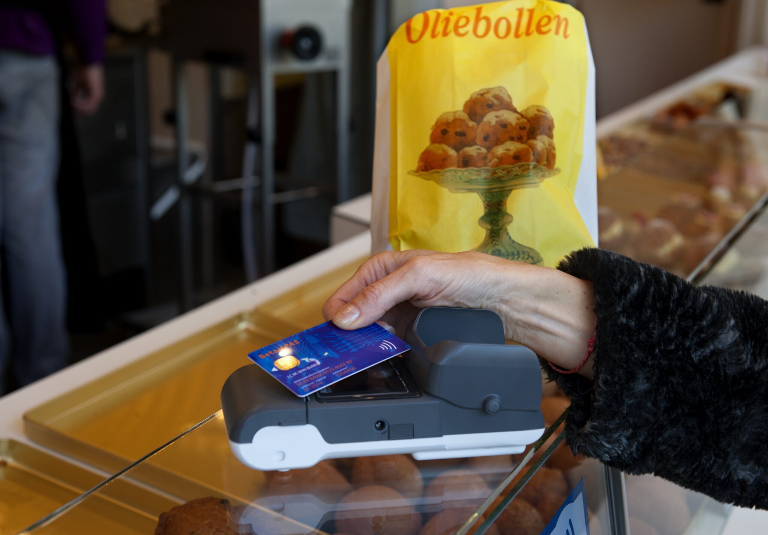 Klant houdt betaalpas tegen pinterminal voor contactloze betaling bij oliebollenkraam, met zak oliebollen op de toonbank.