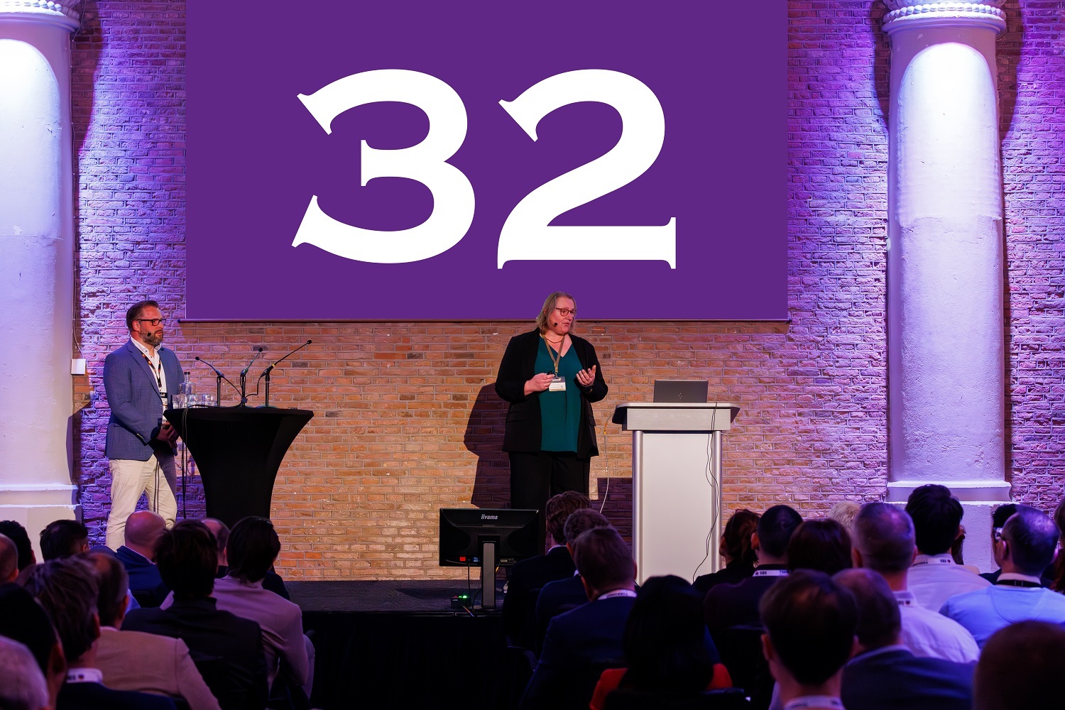 Spreker presenteert op podium tijdens congres, met groot scherm met het getal 32 op de achtergrond en publiek dat aandachtig luistert.