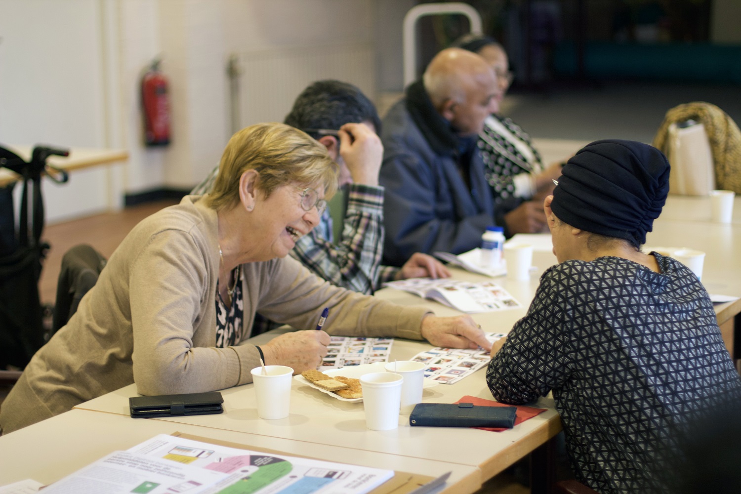 Oudere vrouw helpt een andere deelnemer aan tafel met het invullen en bespreken van papieren, in een rustige groepsbijeenkomst met koffie.