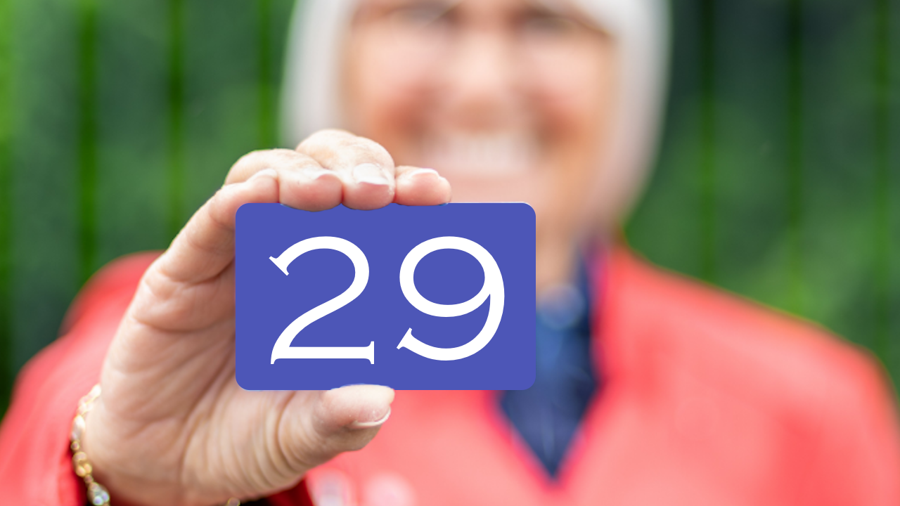 Oudere vrouw houdt een blauw kaartje met het getal 29 naar voren in een groene buitenomgeving, waarbij de focus ligt op het cijfer en haar hand.