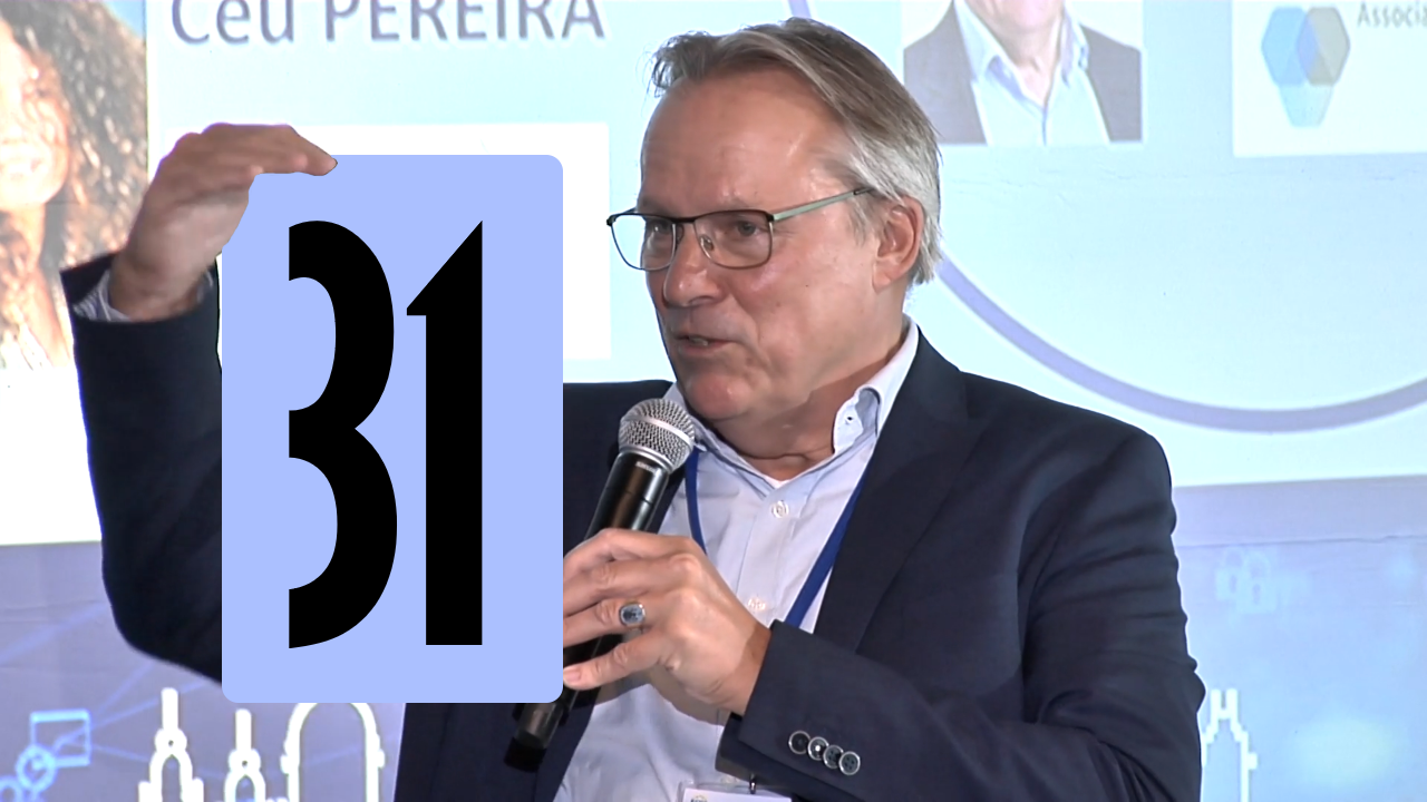 Spreker met microfoon licht een punt toe tijdens een conferentie, met presentatie op de achtergrond en aandachtig publiek buiten beeld.