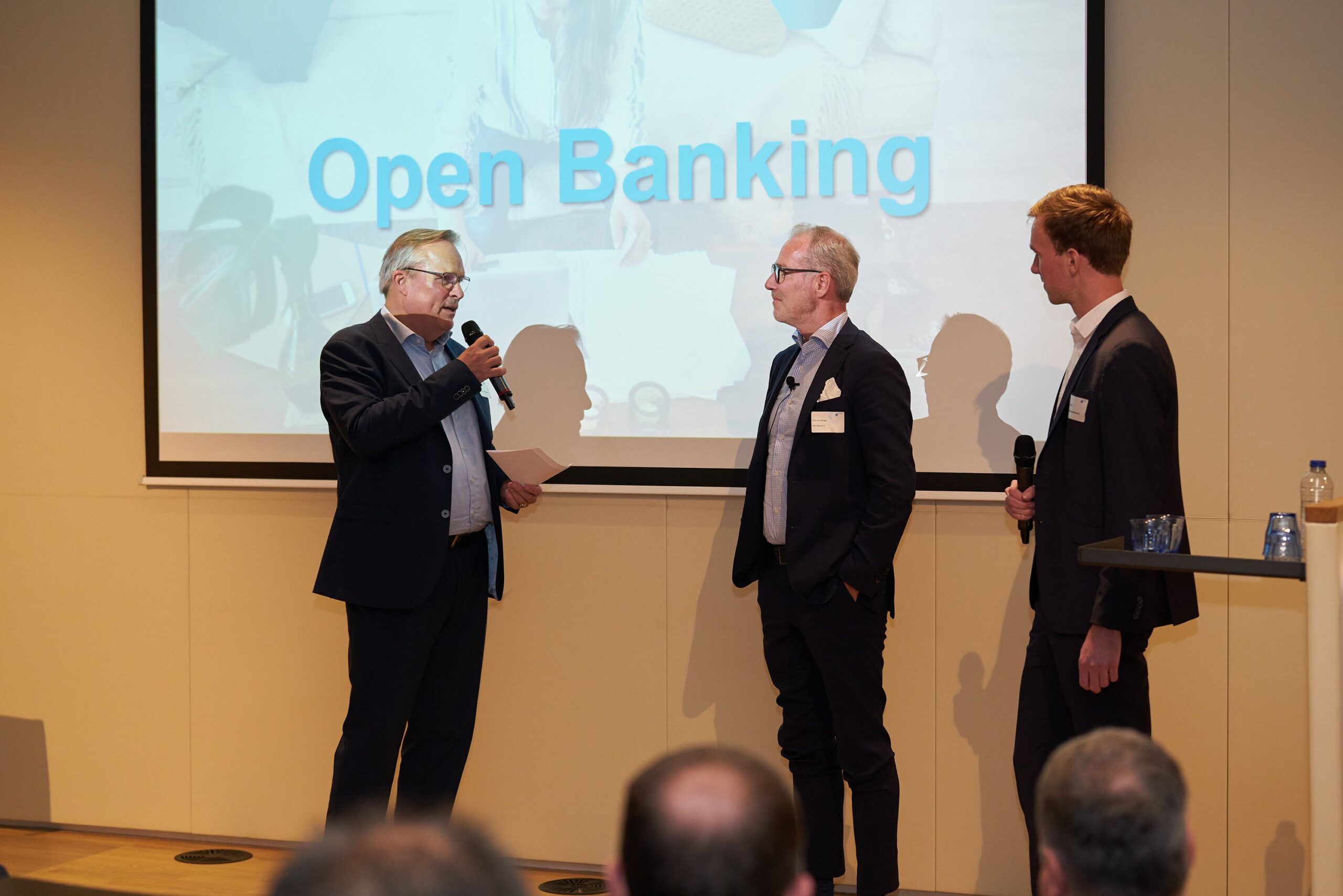 Drie zakelijk geklede sprekers op podium voor scherm met tekst ‘Open Banking’ tijdens panelgesprek voor publiek.