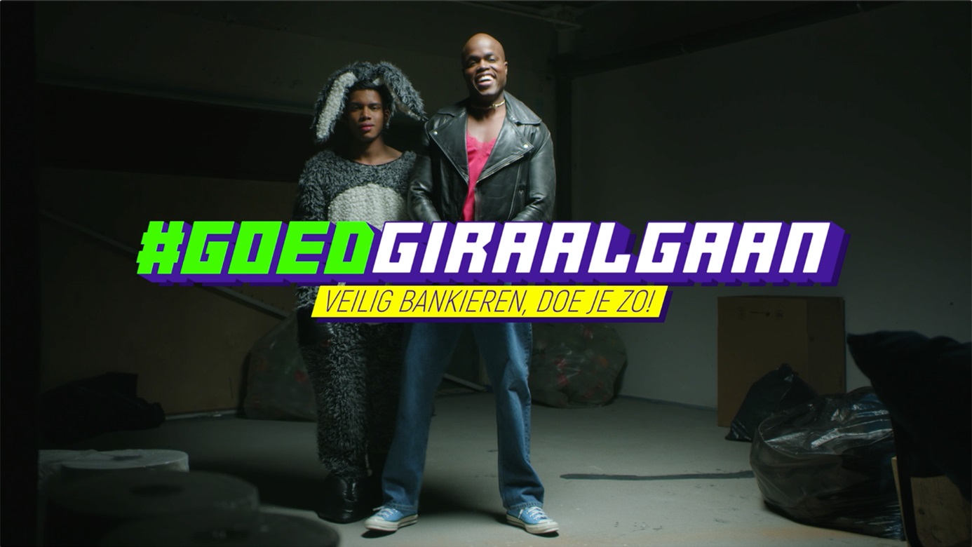 Twee personen staan zelfverzekerd in een industriële ruimte, met de campagneboodschap ‘#GOEDGIRAALGAAN – Veilig bankieren, doe je zo!’ groot in beeld.
