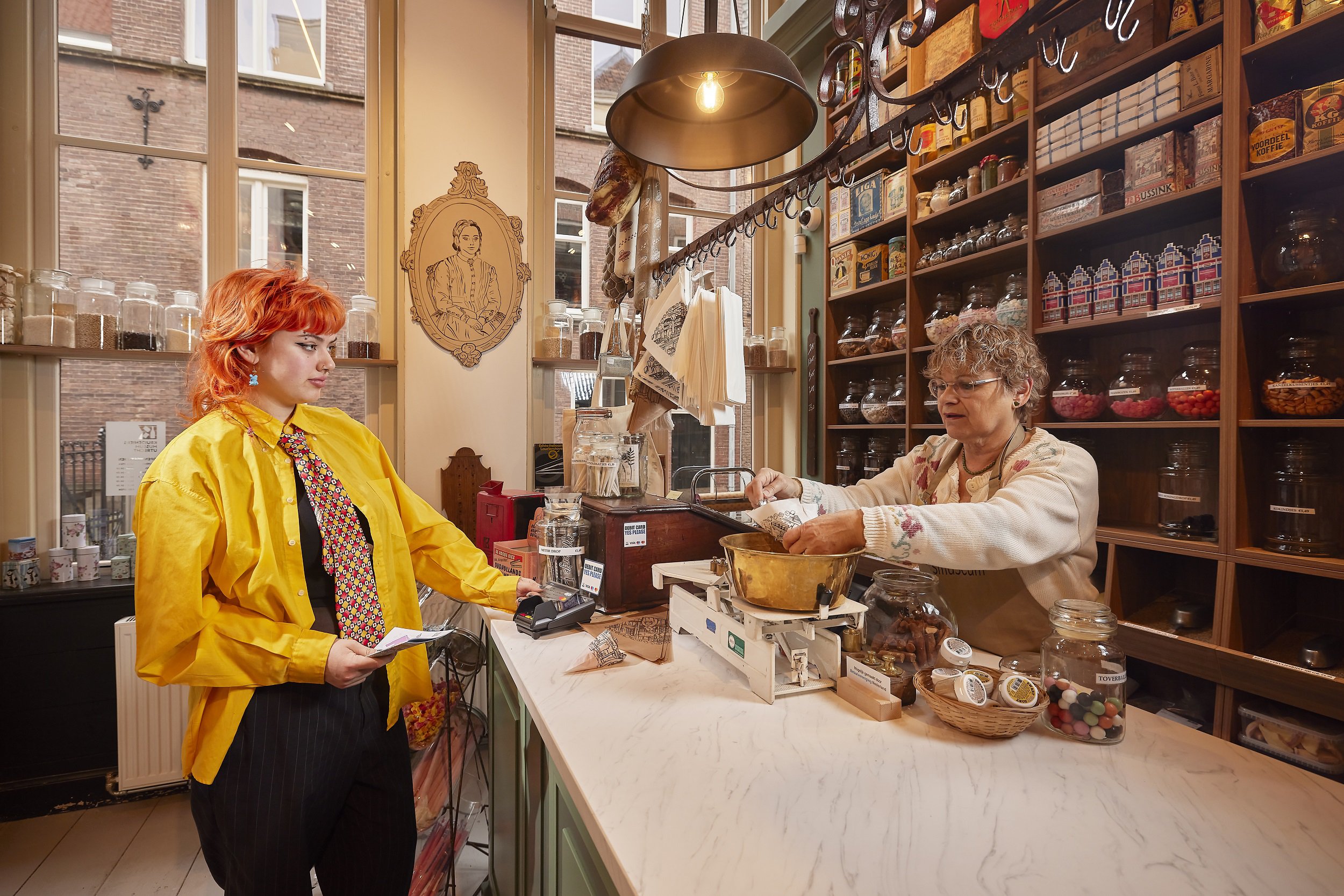 Jonge klant in felgele blouse rekent snoep af in nostalgische snoepwinkel, terwijl de verkoopster snoep weegt achter de toonbank.