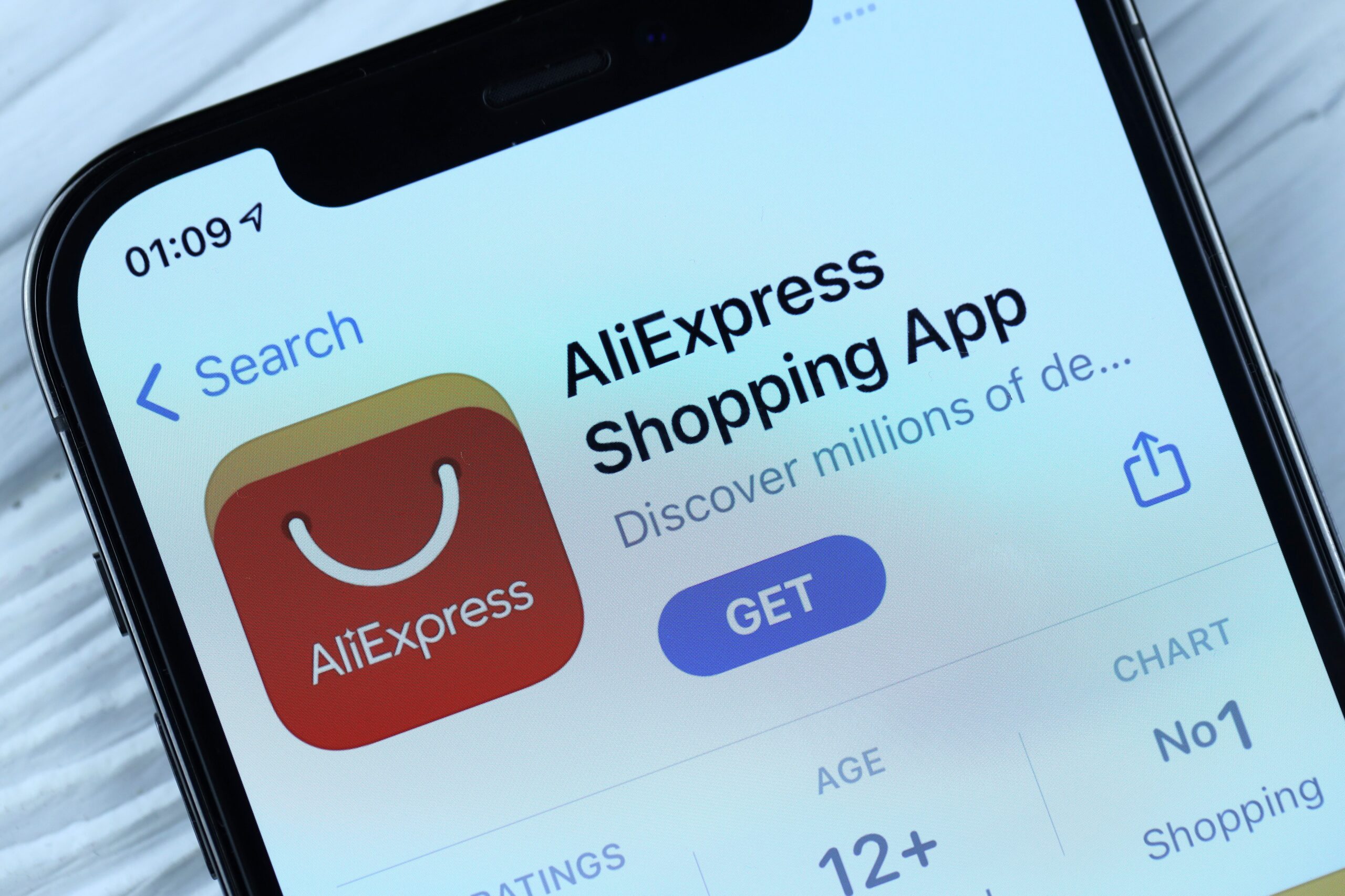 Close-up van een smartphone waarop de downloadpagina van de AliExpress Shopping App in een appstore te zien is. Links staat het rode AliExpress-pictogram met een wit glimlach-tasje, rechts de tekst “AliExpress Shopping App” met daaronder de knop “GET” in blauw en kleine tekst “Discover millions of de…”. Rechts onderin is een deel van de app-gegevens zichtbaar, zoals leeftijdsgrens 12+ en dat de app op nummer 1 in de shopping-hitlijst staat.