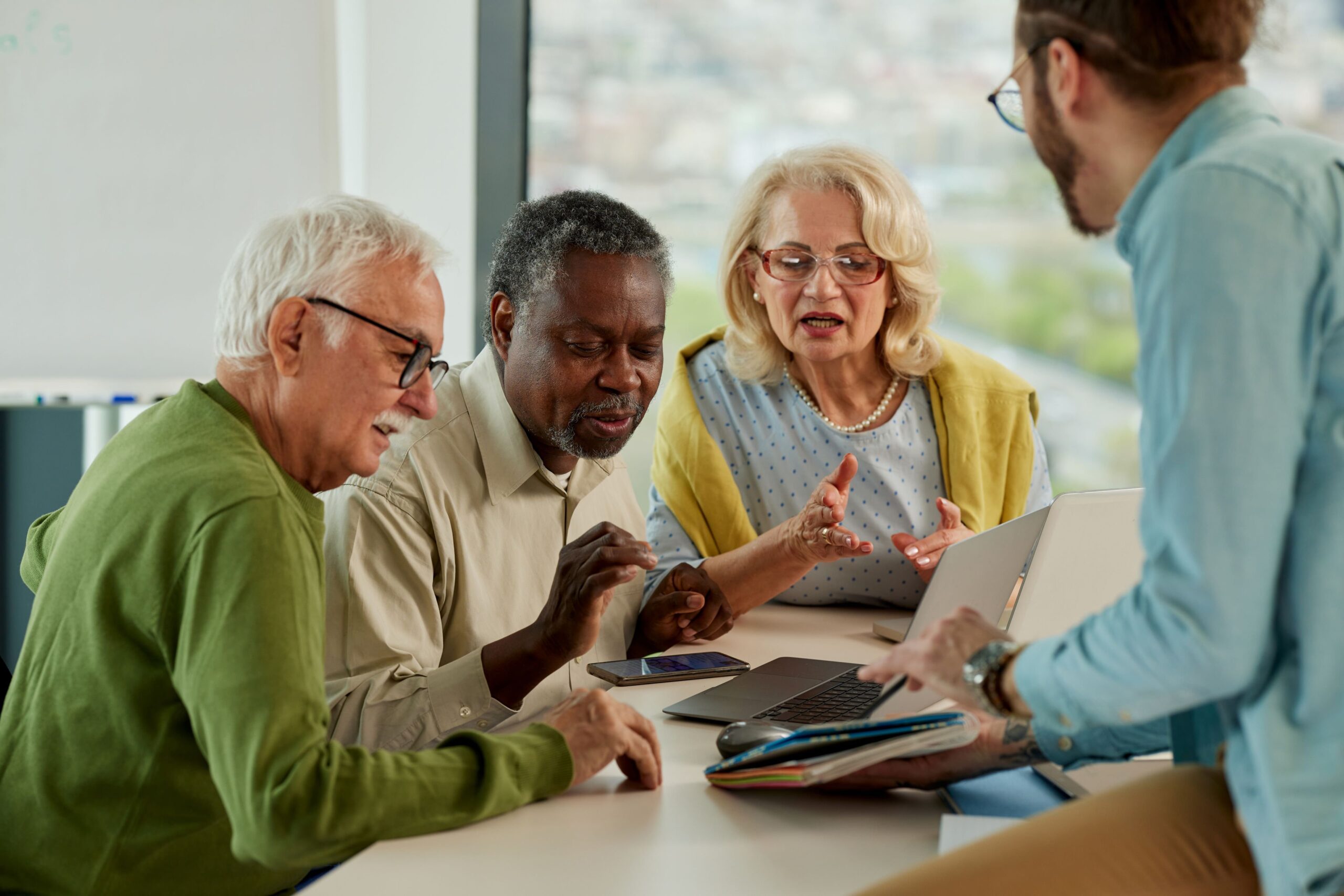 Groep oudere mensen krijgt uitleg van jongere man bij laptop en smartphone tijdens digitale vaardigheidstraining.