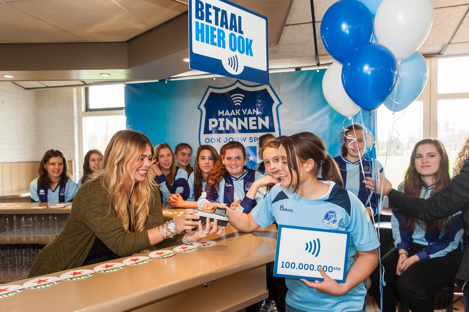 Jonge sporter doet contactloze betaling bij sportkantinebar; team viert de 100 miljoenste transactie met ballonnen.