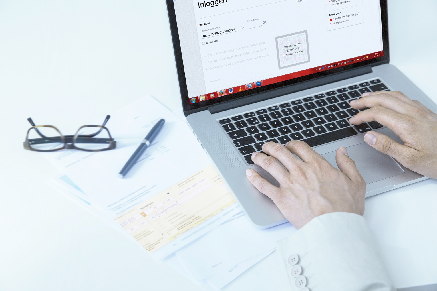 Persoon logt in op internetbankieren op laptop aan wit bureau, met formulieren, acceptgiro, bril en pen naast het toetsenbord.