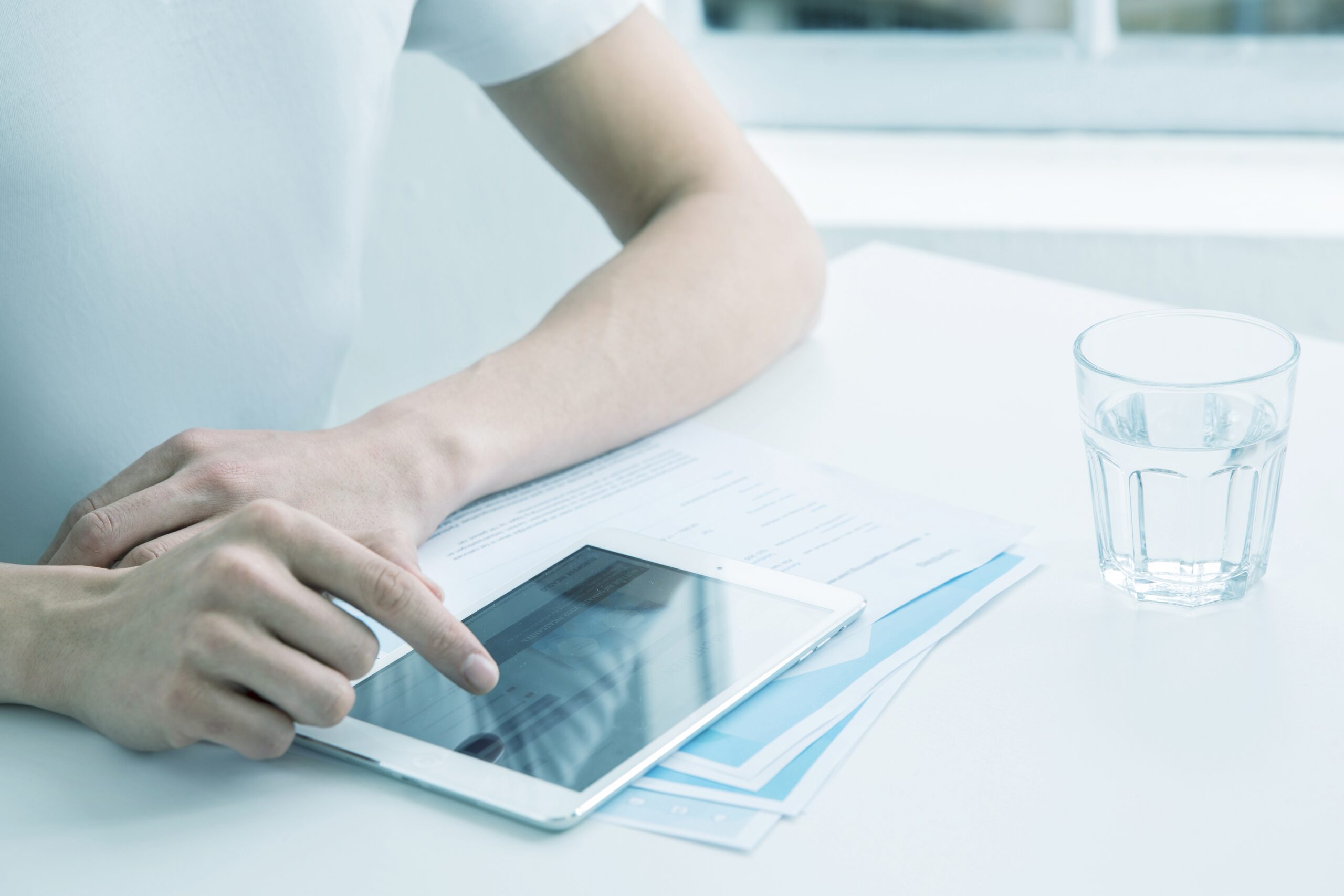 Persoon bedient tablet met digitale documenten op licht bureau, naast papieren en glas water in rustige kantooromgeving.