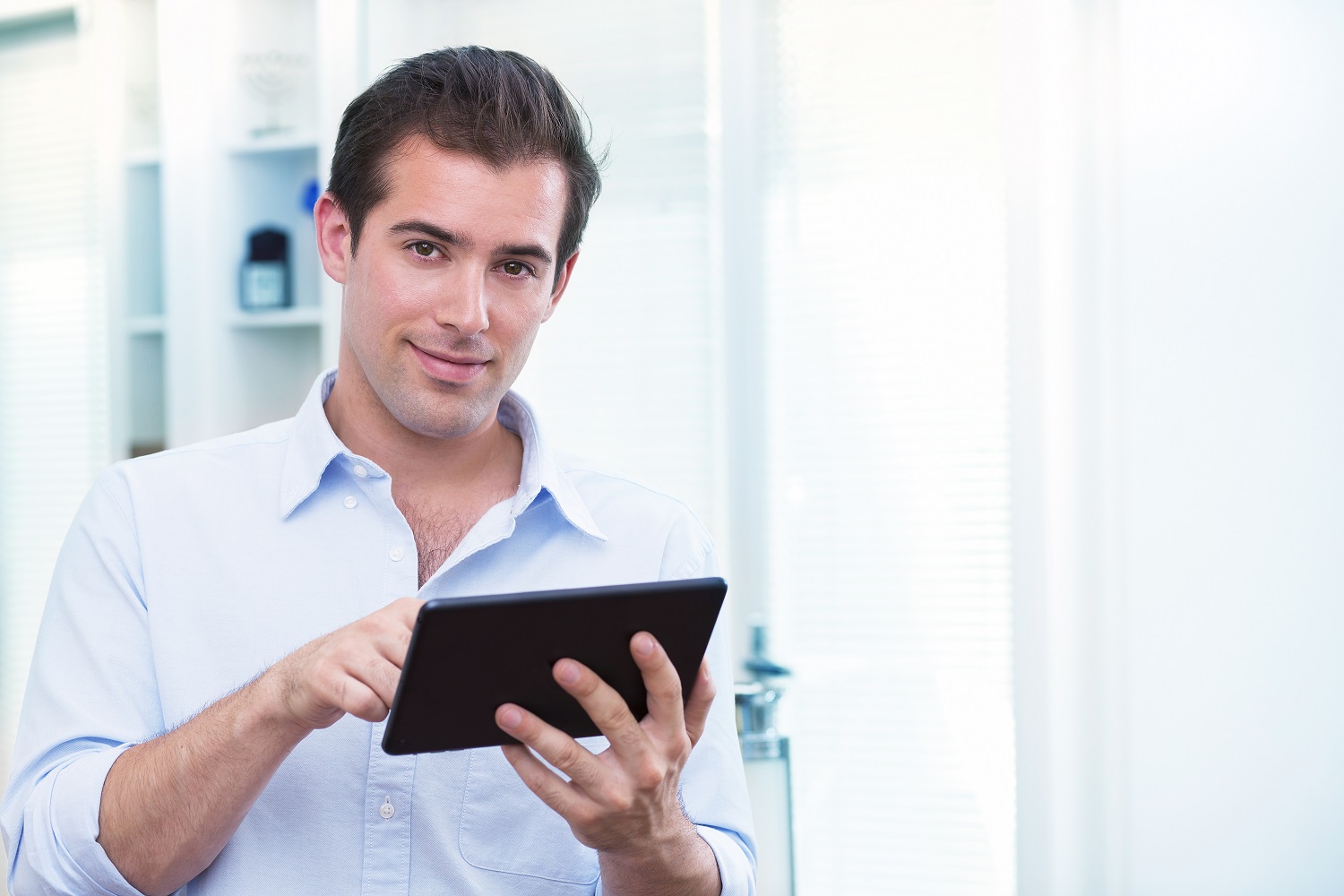 Jonge man in lichtblauw overhemd kijkt glimlachend naar de camera terwijl hij op een tablet tikt in een lichte, moderne omgeving.