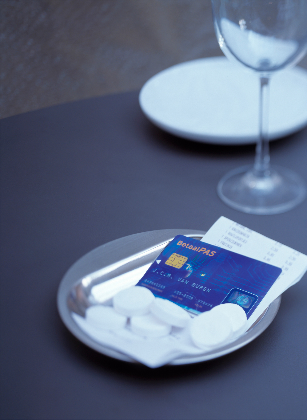 Betaalpas en kassabon liggen op een schoteltje met pepermuntjes op een restauranttafel, naast een wijnglas, als symbool voor afrekenen per pin in de horeca.