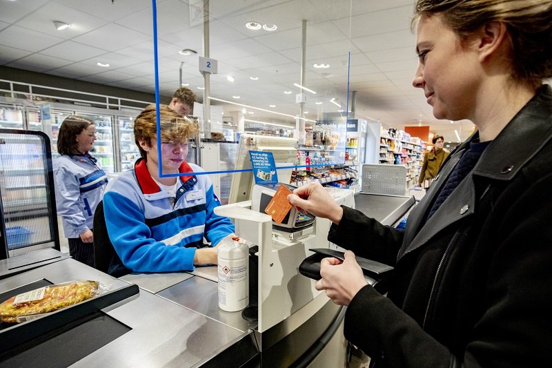 Customer pays at a supermarket counter by presenting a debit card to the cashier, separated by a transparent screen.