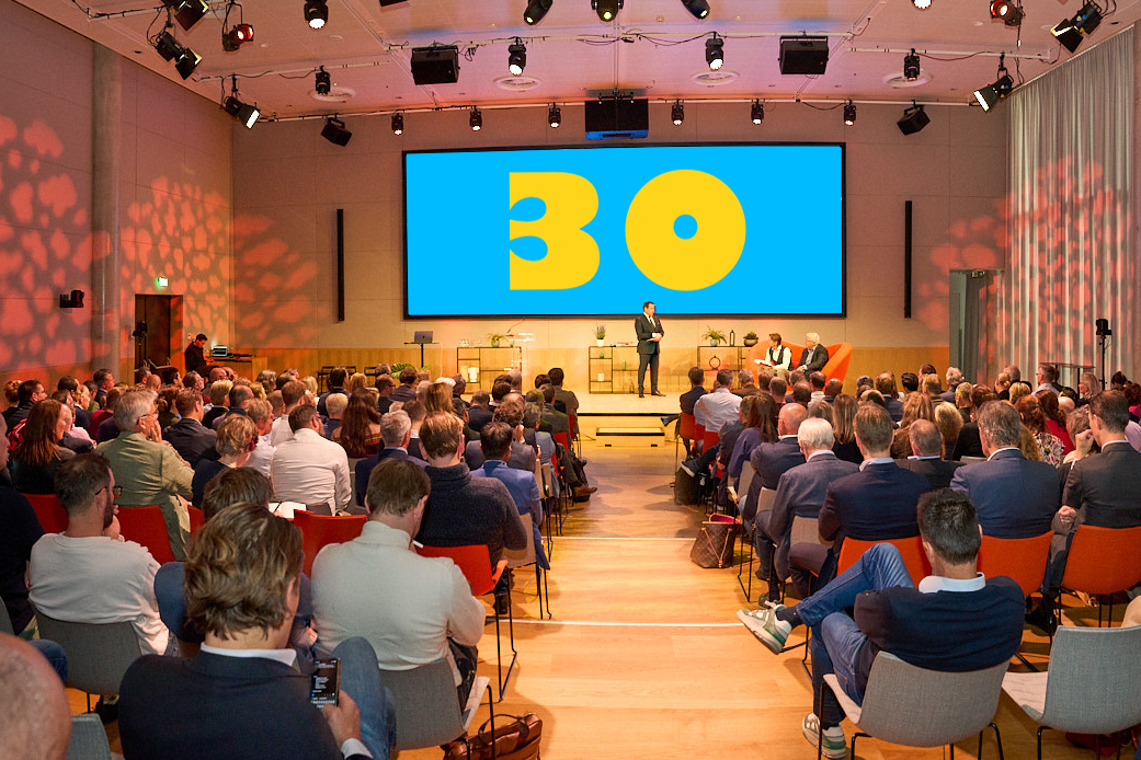 Grote zaal met publiek luistert naar spreker op podium tijdens een professioneel congres, met groot scherm waarop het getal 30 centraal wordt getoond.