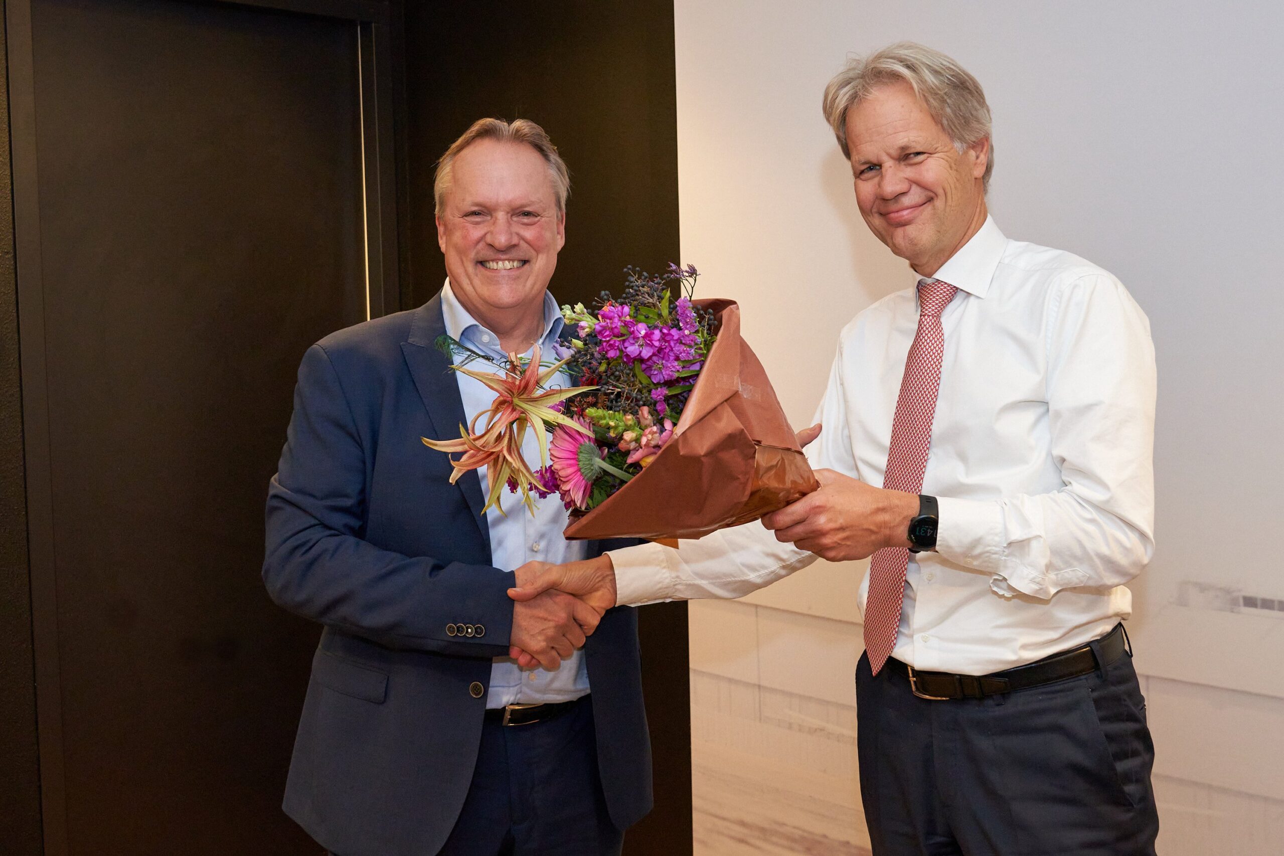 Two men in suits shake hands while smiling while a bouquet of flowers is presented upon the appointment of the new director the Dutch Payments Association.