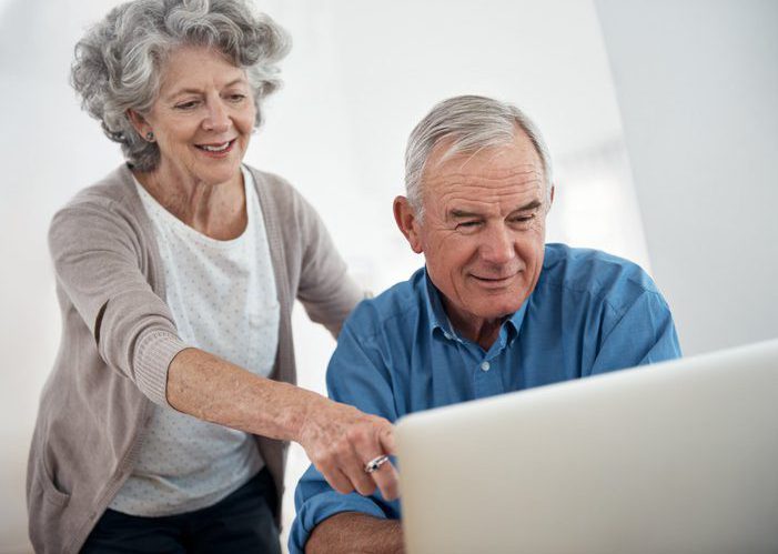 Older couple sits together at a laptop while the woman smiles and points at something on the screen in a bright home environment.