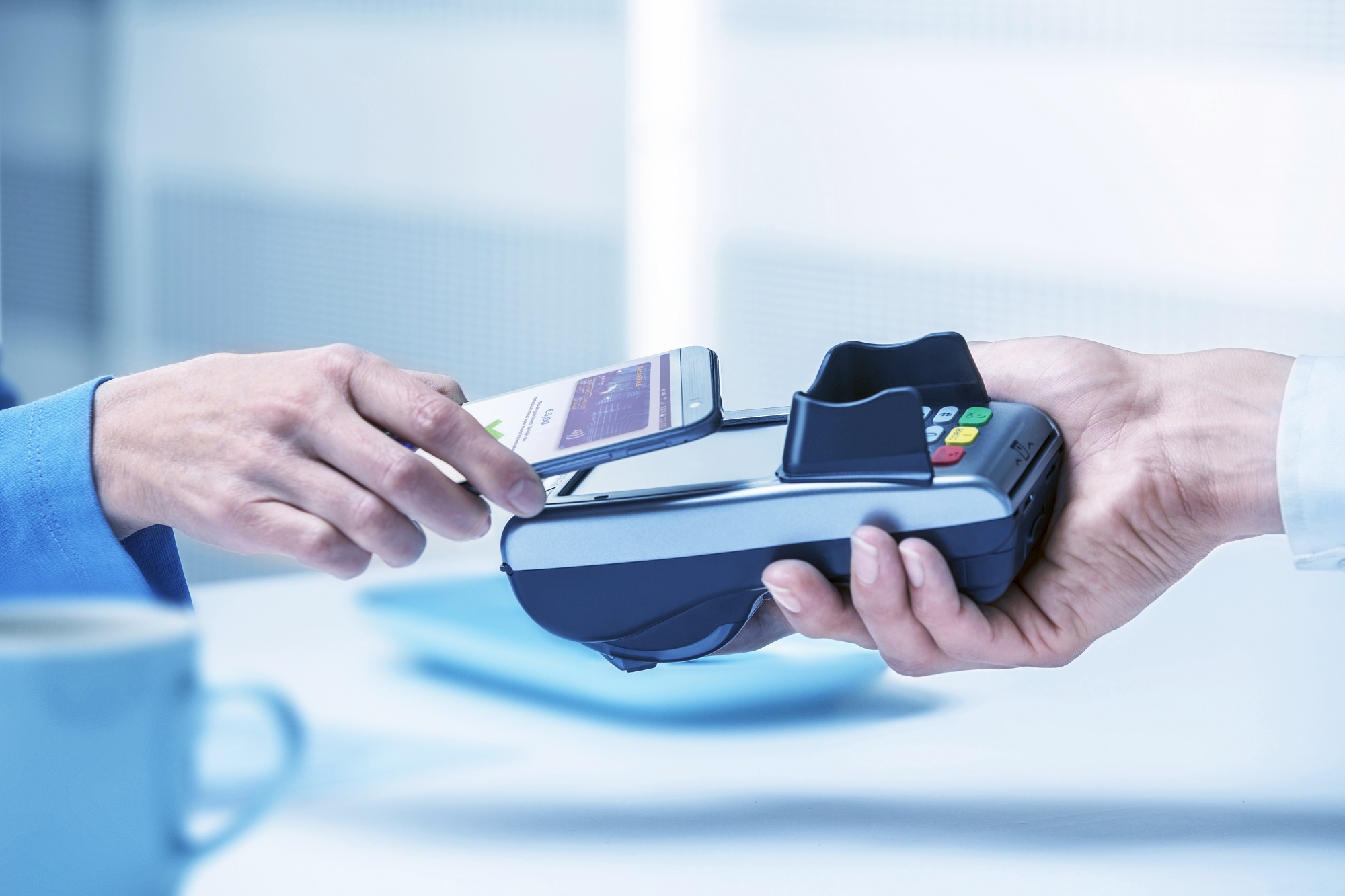 Hand holds a smartphone against a payment terminal for a contactless payment, while an employee holds the device in a professional environment.