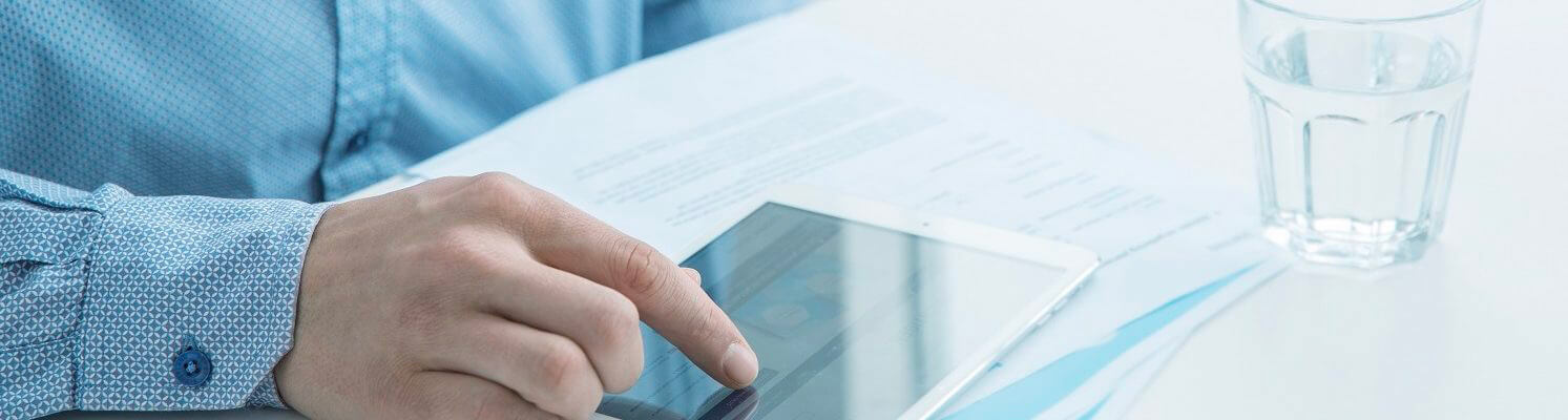 Hand of person in blue shirt operates tablet above financial documents on desk with glass of water.
