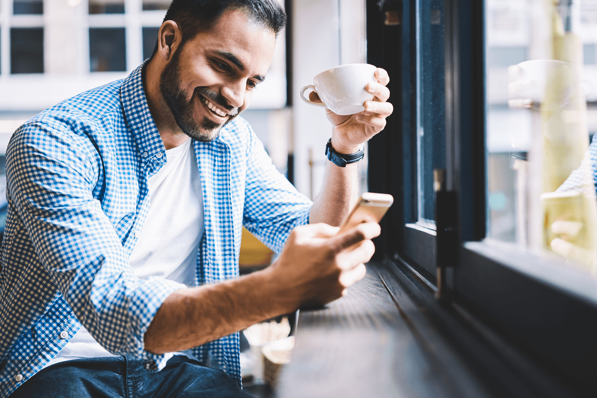 Man in a coffee shop laughs while arranging something via a mobile app with his smartphone and cup of coffee by the window.