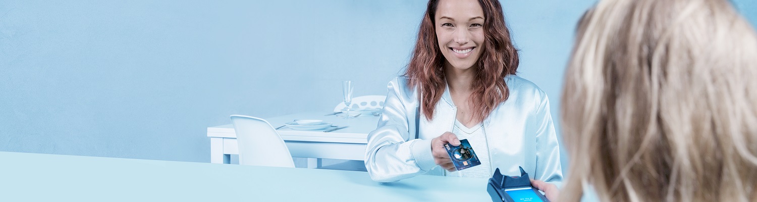 Woman smilingly hands over her debit card to an employee holding a POS terminal during a leisurely contactless payment at the table.