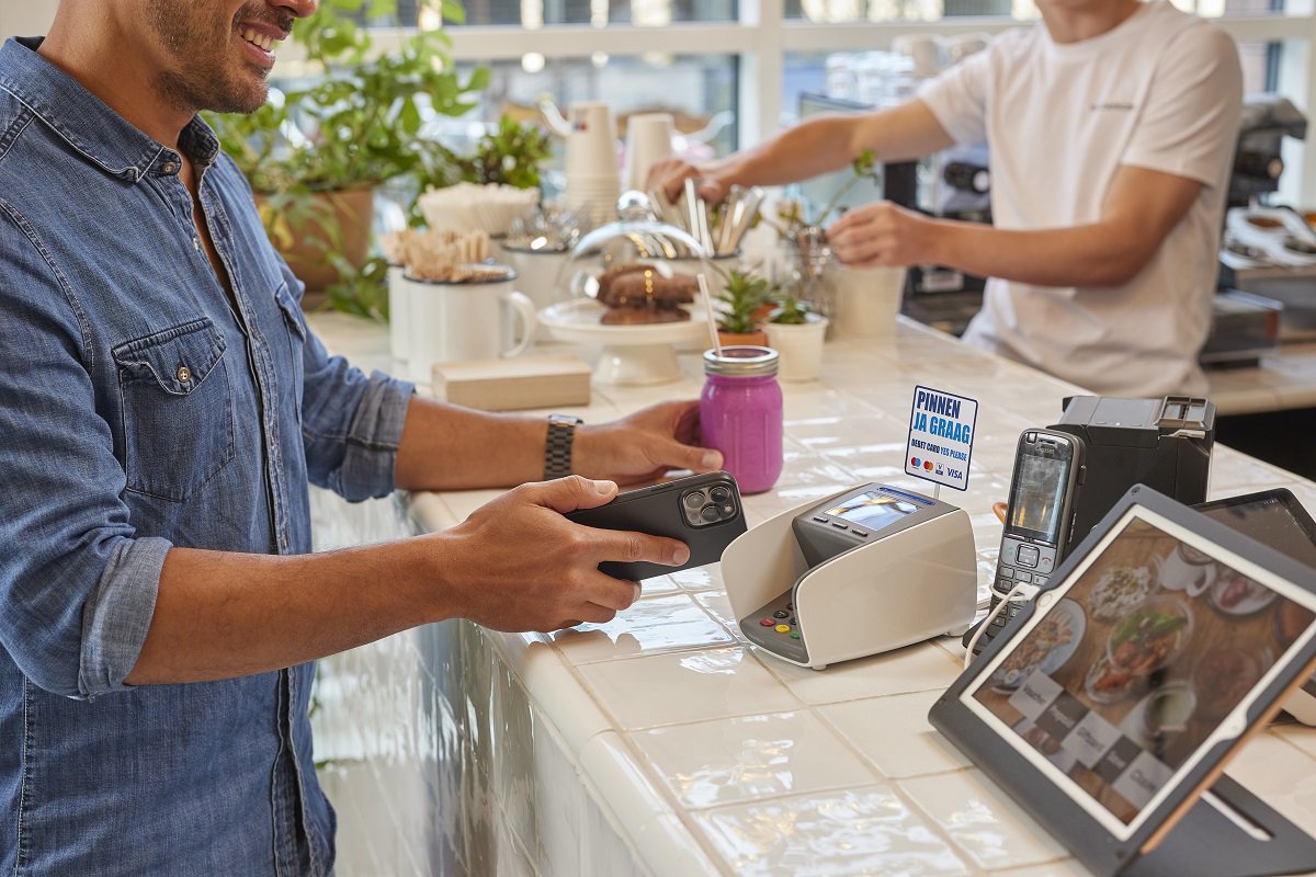 Smiling man makes contactless payment with smartphone at coffee bar, while barista prepares drinks in the background.