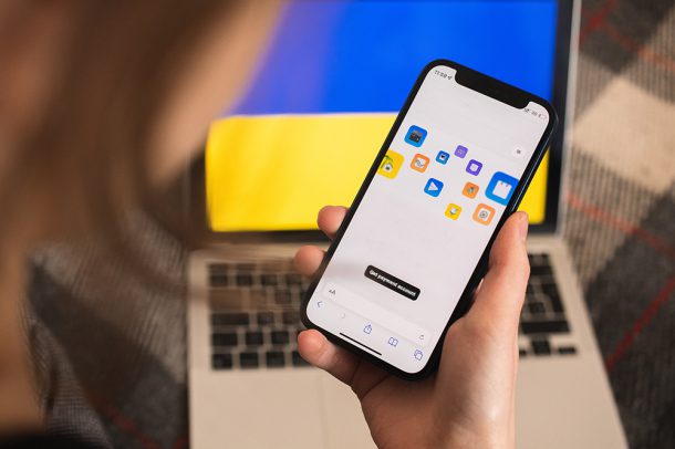 Person holds a smartphone displaying a payment account application in front of a laptop showing the Ukrainian flag.