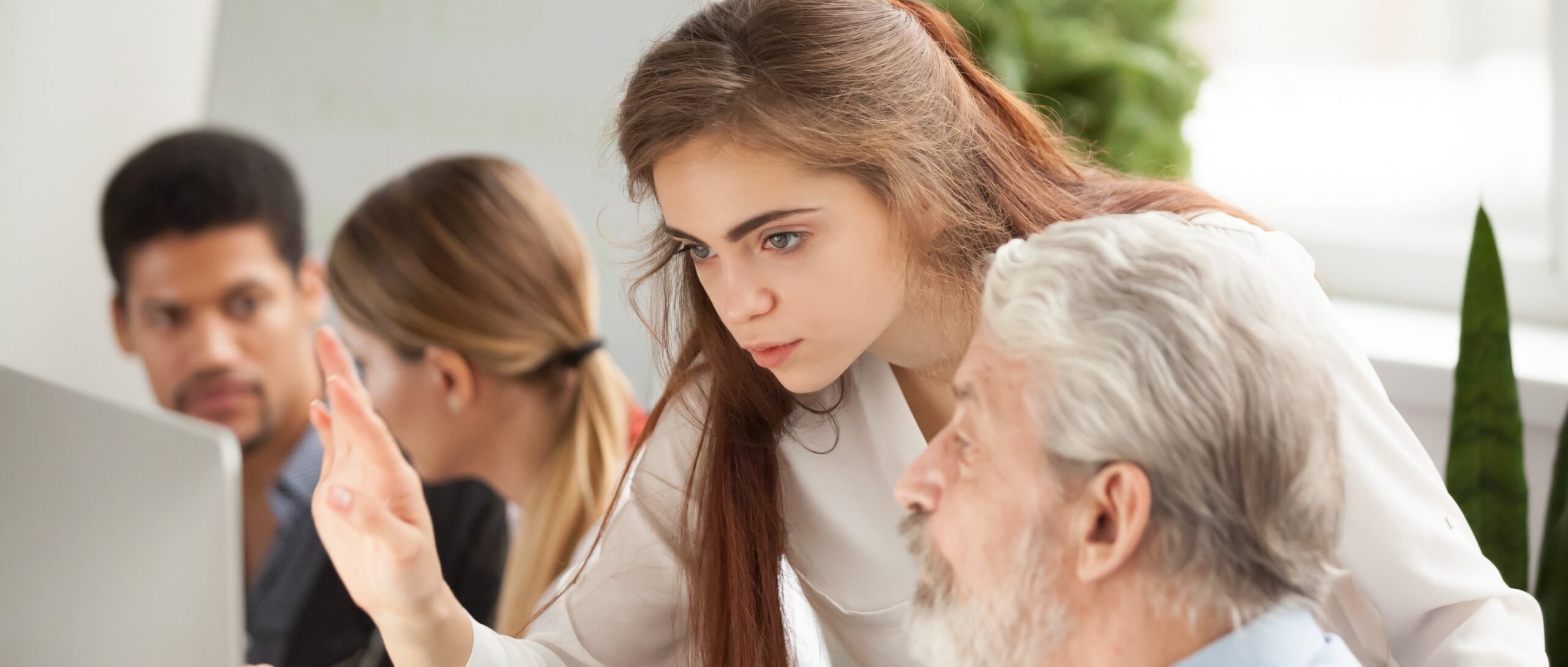 Employee calmly explains digital banking environment to older colleague at desk, while team works in the background in bright office space.