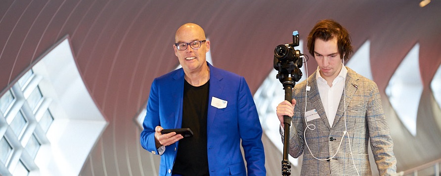 Presenter Vincent Everts with a tablet and cameraman Jelle Crop with a video camera on a tripod walk through a modern, bright hall during an event recording.