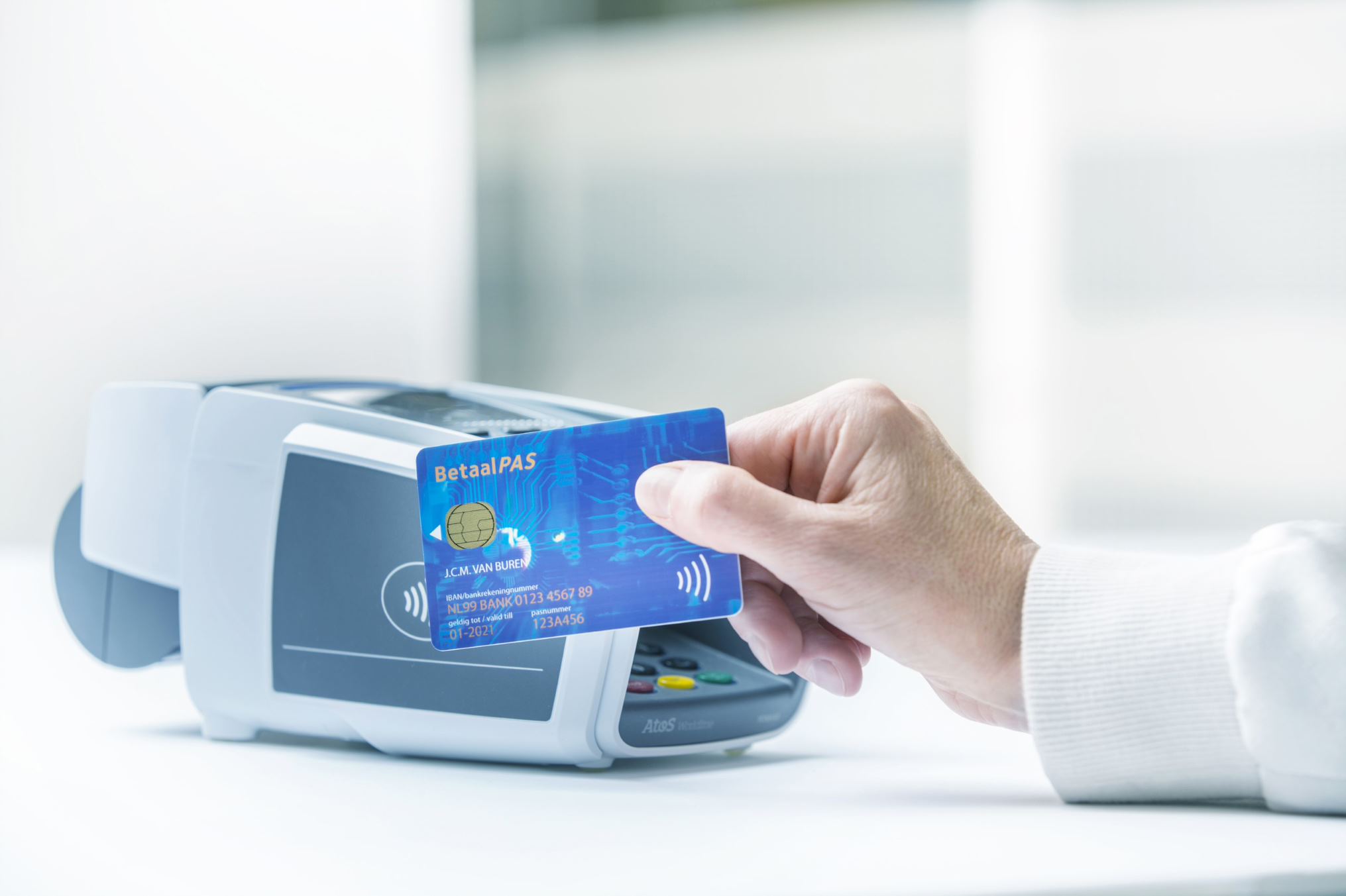 Hand holds a debit card against a POS terminal for a contactless payment in a bright, professional retail or office environment.