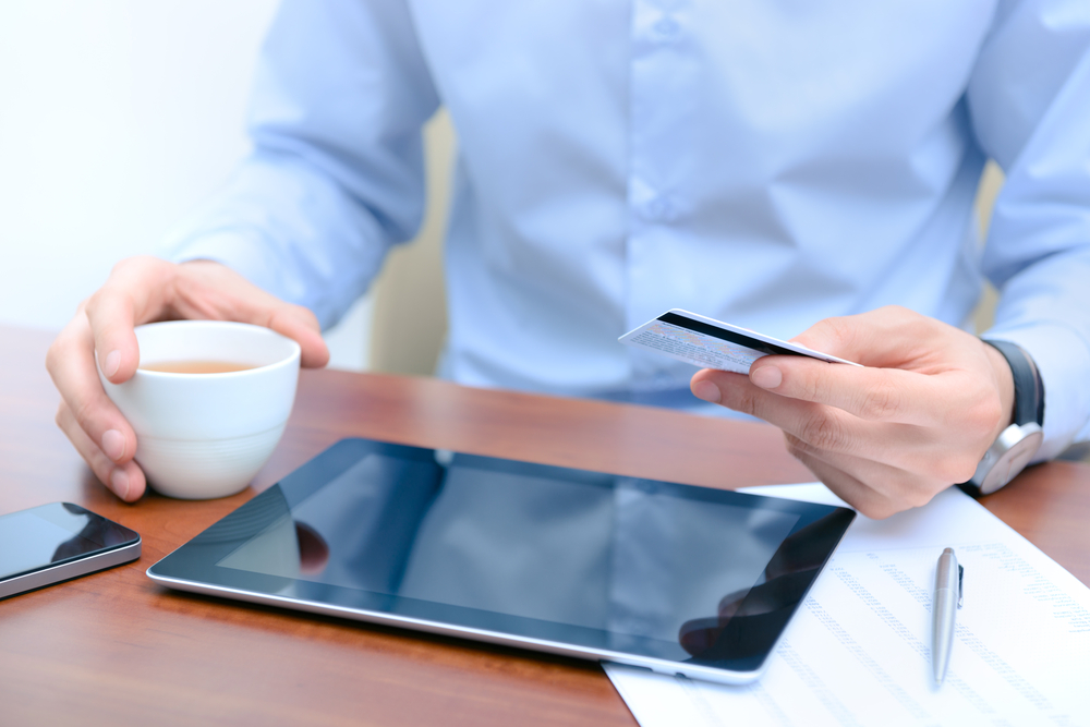 Office worker with debit card in hand is looking at tablet for online payment, with coffee, smartphone and documents on the desk.