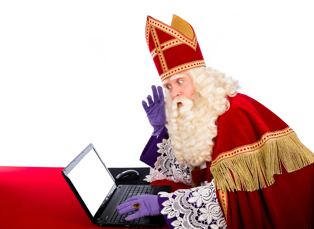Sinterklaas sits behind a laptop on a red tablecloth and looks at the screen in surprise in a bright studio.