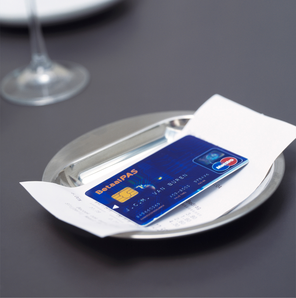Blue debit card lies on a metal tray on a set table together with a restaurant bill.