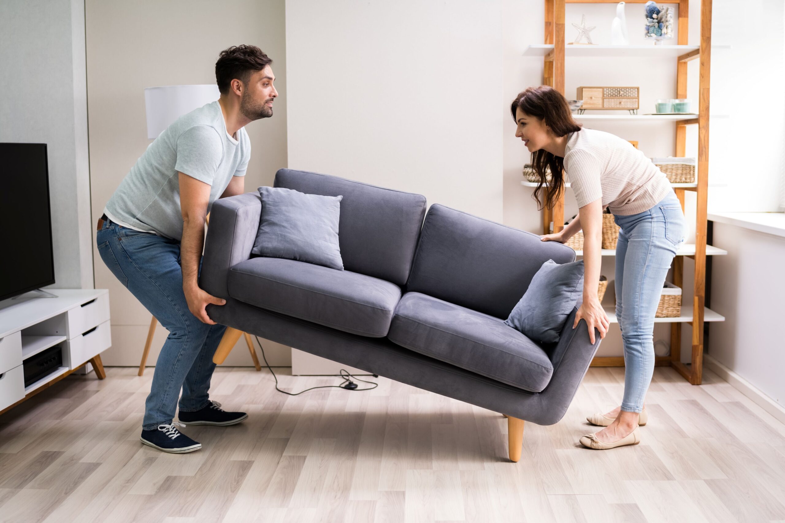 Man en vrouw tillen samen een grijze bank in een lichte woonkamer, terwijl ze lachend de nieuwe opstelling bepalen.