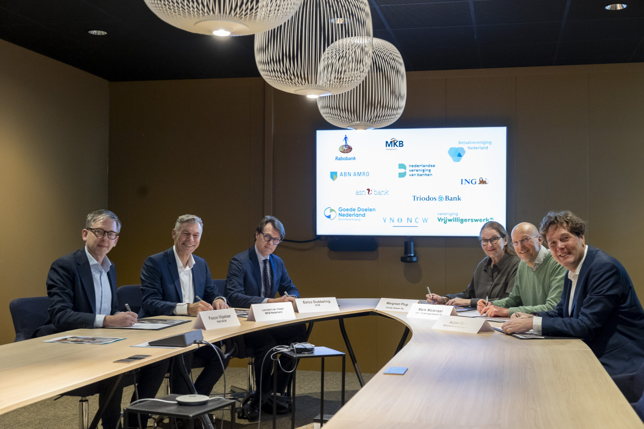 Bestuurders van banken en organisaties, waaronder Betaalvereniging Nederland, ondertekenen gezamenlijk een samenwerking aan een vergadertafel.