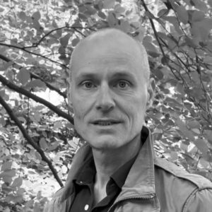 Zwart-witportret van een kale man in jas, buiten tussen bladeren en takken, die rustig in de camera kijkt.