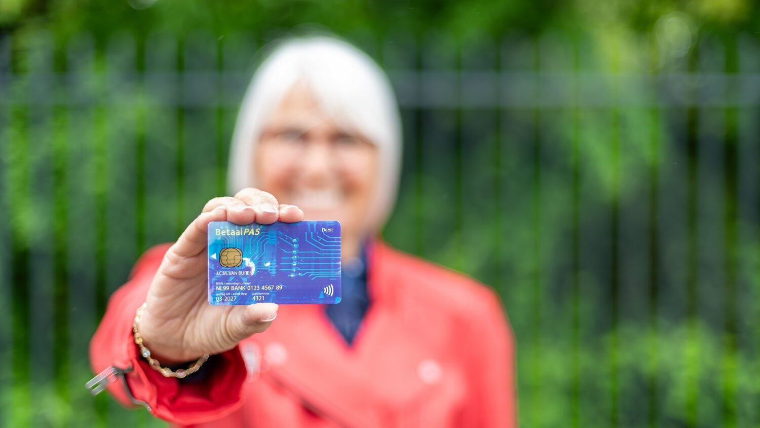 Oudere vrouw houdt een betaalpas duidelijk naar voren in een groene buitenomgeving, waarmee zij laat zien dat zij eenvoudig en zelfstandig kan betalen.