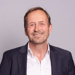 A man smiles warmly in front of a yellow background in a bright and approachable business portrait.