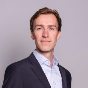 A man in a suit looks calmly at the camera against a dark background, giving a professional impression.