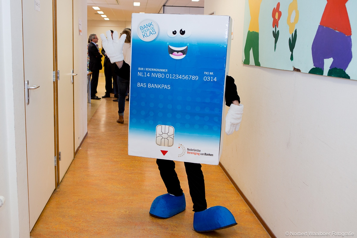 Mascotte Bas Bankpas loopt door een schoolgang: een persoon in een groot blauw bankpas-kostuum met lachend gezicht, witte handschoenen en blauwe schoenen, onderdeel van de educatieve campagne Bank voor de Klas.