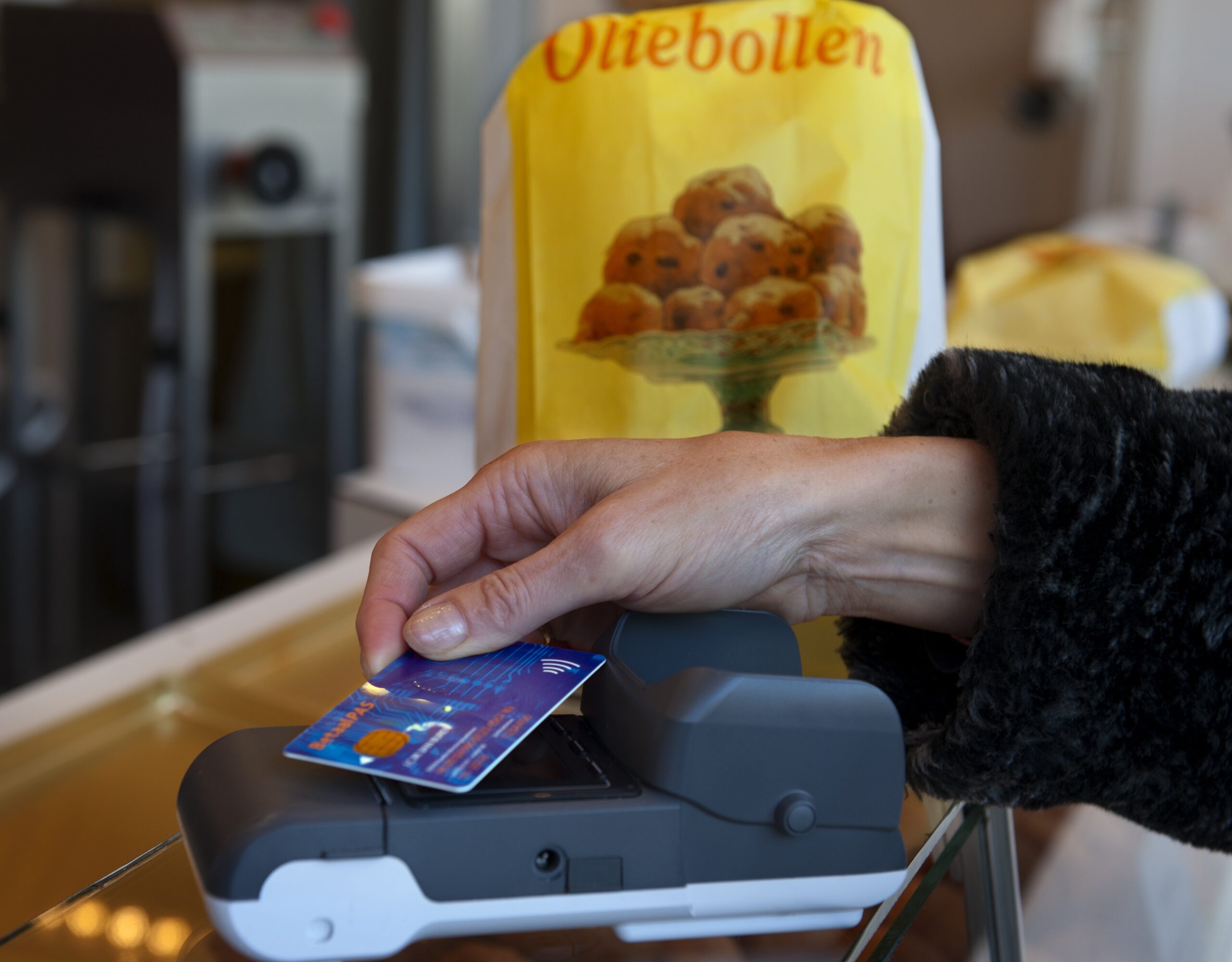 Hand houdt betaalpas contactloos tegen pinterminal bij oliebollenkraam, met gele zak oliebollen op de achtergrond.