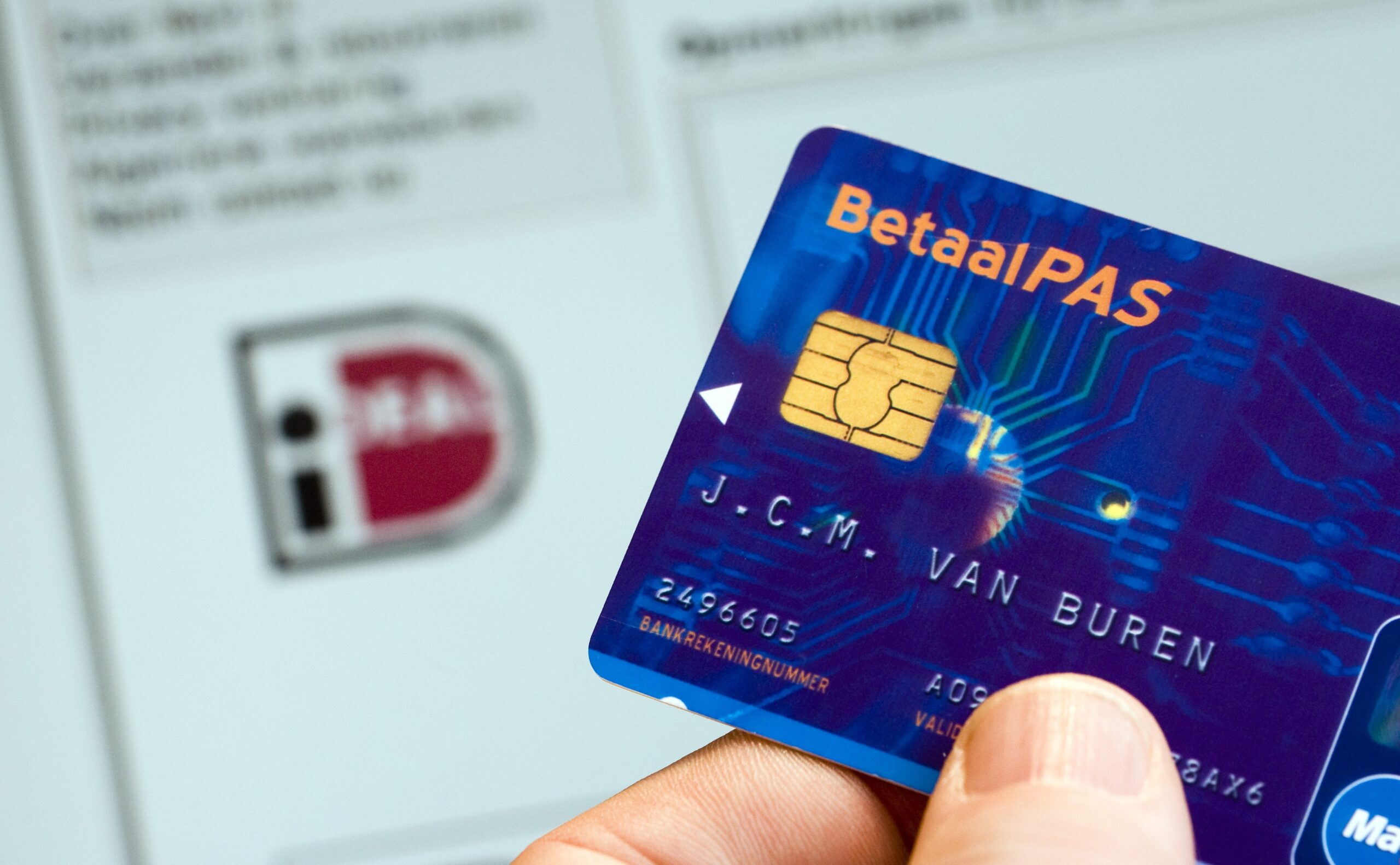 Hand houdt blauwe betaalpas met chip voor scherm met iDEAL-logo, symbool voor veilig online afrekenen via bankrekening.