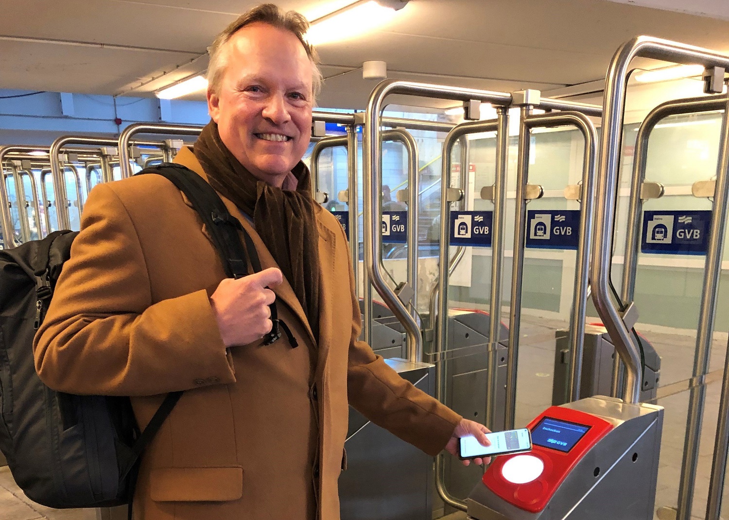 Man in camelkleurige jas checkt in bij GVB-poortje met zijn smartphone voor contactloos reizen in het openbaar vervoer.