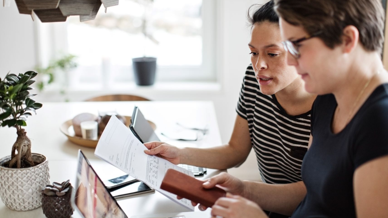 Twee vrouwen bekijken samen rekeningen en een portemonnee aan de keukentafel, terwijl ze betalingen en uitgaven bespreken met laptop en papieren.
