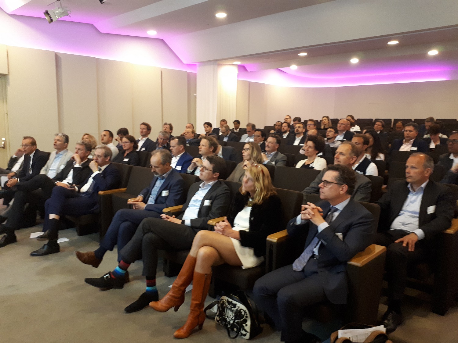 Zaal vol zakelijke bezoekers luistert aandachtig naar presentatie in auditorium met zachte paarse sfeerverlichting.
