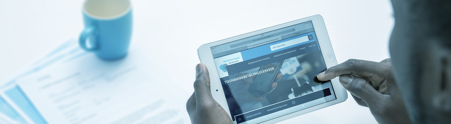 Persoon bekijkt op een tablet de website over toonbankbetalingsverkeer van Betaalvereniging Nederland, liggend op een bureau met documenten en een koffiemok.