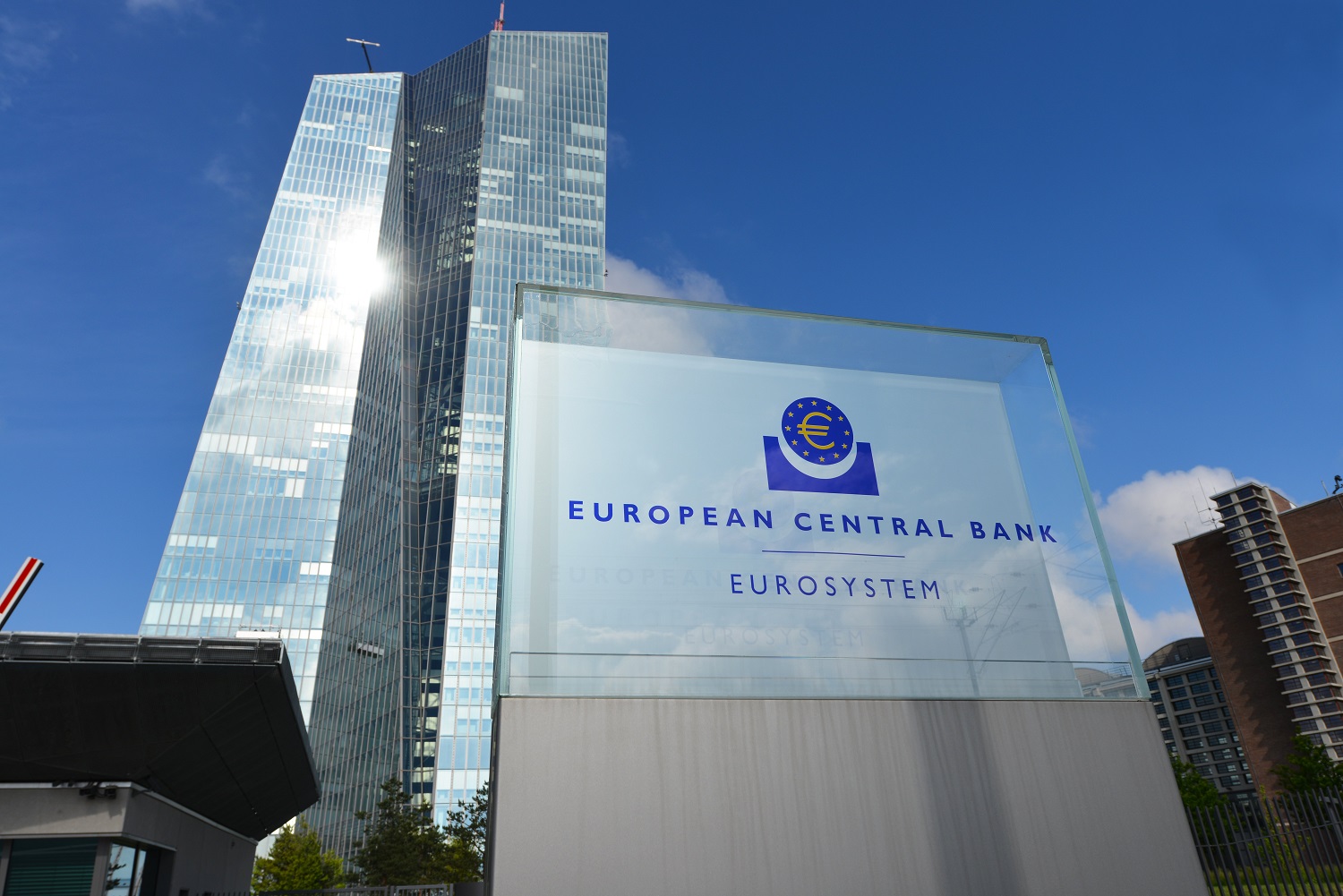 Glazen hoofdkantoor van de Europese Centrale Bank met groot ECB-logo op voorgrond tegen blauwe lucht.