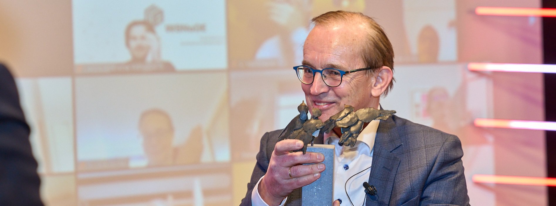 Piet Mallekoote houdt glimlachend een beeldje vast op een podium, tijdens een uitreiking of evenement in een professionele setting.