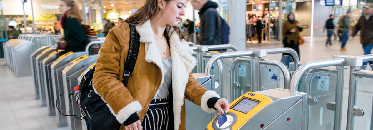 Jonge vrouw checkt in met OV-chipkaart bij NS-toegangspoortje op druk station, met andere reizigers op de achtergrond.