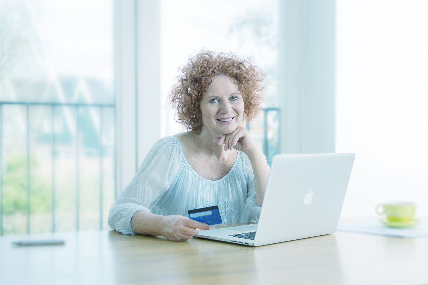 Vrouw met krullend rood haar zit glimlachend aan een houten tafel met een open laptop voor zich. In haar hand houdt ze een betaalpas of creditcard vast, alsof ze online een aankoop of bankhandeling doet. Op de achtergrond zijn grote ramen met licht en een zachte, lichte woonkamer te zien.
