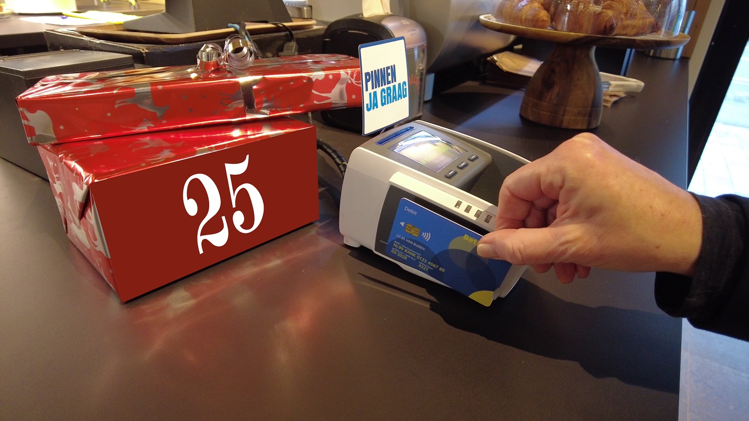 Hand houdt betaalpas tegen pinterminal voor contactloze betaling naast rood ingepakt cadeau met nummer 25 en bordje ‘Pinnen ja graag’.