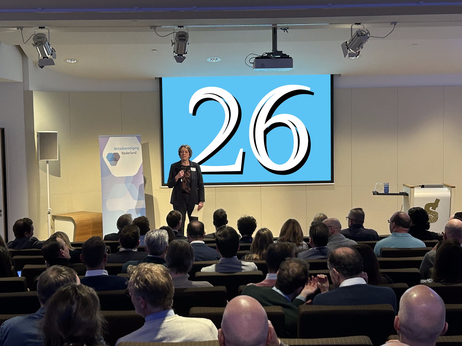 Spreker van Betaalvereniging Nederland presenteert voor volle zaal, met groot wit cijfer 26 op blauw projectiescherm.